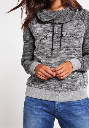 Femme portant un sweat-shirt gris marbré avec "true" en écriture noire et des poignets gris clair, associé à un jean en denim bleu.