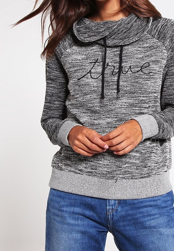 Femme portant un sweat-shirt gris marbré avec "true" en écriture noire et des poignets gris clair, associé à un jean en denim bleu.