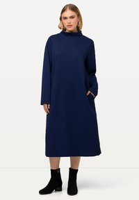 Marineblaues Langarmkleid mit hohem Kragen, strukturiertem Muster, oversized Schnitt und seitlichen Taschen. Kombiniert mit schwarzen Knöchelstiefeln.