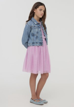 Giacca di jeans con dettagli in borchie, abito in tulle rosa a strati e décolleté blu. L'outfit presenta un design casual ma stratificato.