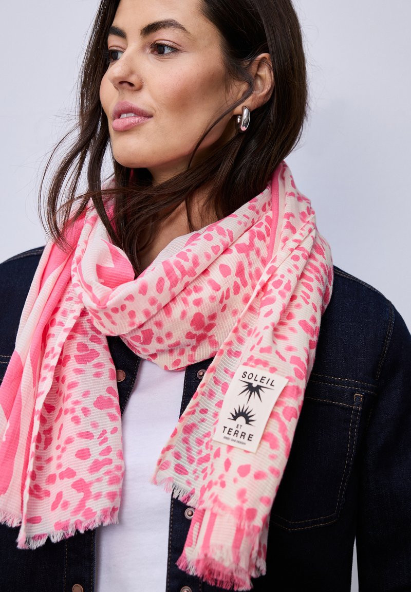 Femme portant des boucles d'oreilles en argent, une veste en jean, un haut blanc et une écharpe rose et blanche à motif avec une étiquette "Soleil et Terre".