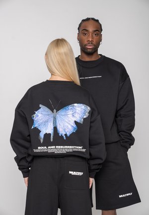 Zwei Personen tragen schwarze Sweatshirts; eine blickt nach vorne, die andere zeigt auf dem Rücken einen großen blauen Schmetterling und den Schriftzug „Seele und Auferstehung“.