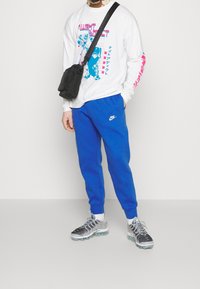 Valkoinen pitkähihainen paita, jossa värikäs grafiikka, siniset joggers-housut ja musta olkalaukku. Hopeanväriset ja mustat kuvioidut lenkkarit läpinäkyvillä pohjilla.