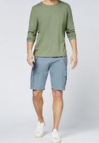 Chemise à manches longues vert olive associée à un short cargo bleu clair avec poches latérales, portée avec des baskets blanches. Design simple et fonctionnel.