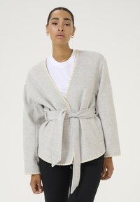 Giacca avvolgente grigio chiaro con cintura, bordi a ondina e tasche laterali, realizzata in materiale morbido; sovrapposta a una t-shirt bianca.