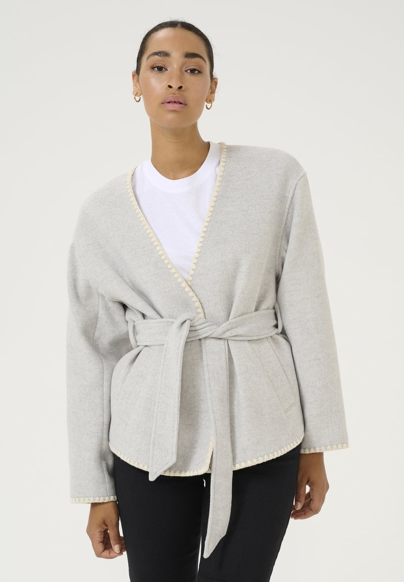 Giacca avvolgente grigio chiaro con cintura, bordi a ondina e tasche laterali, realizzata in materiale morbido; sovrapposta a una t-shirt bianca.