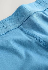Sous-vêtements en coton bleu clair avec une ceinture texturée portant le mot "NEXT" en relief. Surface lisse, coutures discrètes le long des bords.