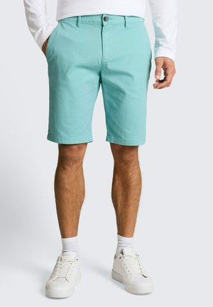 Man met turquoise knielange shorts, wit shirt met lange mouwen, witte sokken en witte sneakers, staand met ontspannen handen.