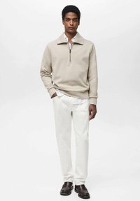 Felpa beige con zip abbinata a pantaloni bianchi e scarpe marroni scure. Presenta un colletto alto e una vestibilità rilassata, realizzata in materiale morbido.