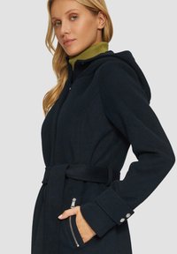 Manteau bleu marine avec capuche, taille ceinturée et poche zippée sur le côté. Fabriqué dans un matériau texturé, il présente des accents de boutons sur les manches.