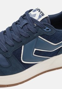 Baskets bleues avec des panneaux en daim et cuir, design en chevron blanc et bleu clair, lacets bleu marine, languette avec logo Levi's, et semelle blanche avec semelle extérieure marron.