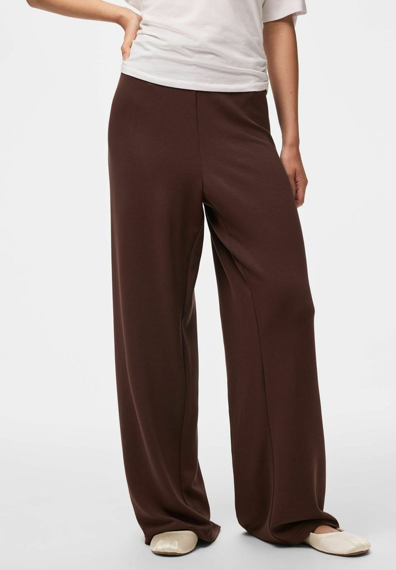 Pantalon ample brun foncé porté avec un haut blanc et des chaussures beige à enfiler sur une personne debout devant un fond uni.