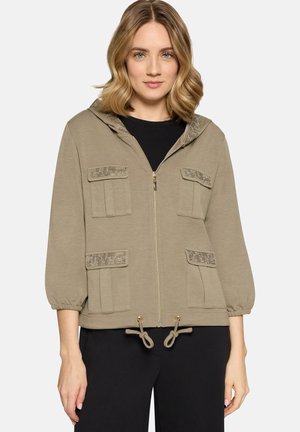 Kvinde iført beige lynlåstrøje med fire forlommer og snore i kanten, kombineret med sorte bukser.