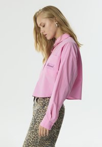 Roze cropped shirt met een spitse kraag, één borstzak en geborduurd detail. Draag het met dierenprint broeken, die een gladde textuur tonen.