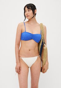 Blå mønstret bikinitopp med tynne stropper, kombinert med en kremfarget bunn. Modell som holder en flerfarget strikket veske.