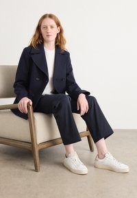 Veste bleu marine à double boutonnage, chemise blanche, pantalon sombre et baskets blanches. Assis sur une chaise beige, design minimaliste, textures lisses.