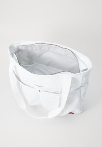 White canvas tote bag featuring a spacious interior, zipper closure, and exterior pocket with a snap button. Sturdy fabric handles.