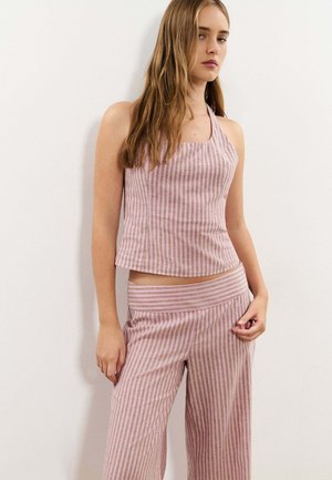 Jeune femme portant un haut sans manches à rayures verticales roses et beiges et un pantalon large assorti, debout contre un mur blanc.