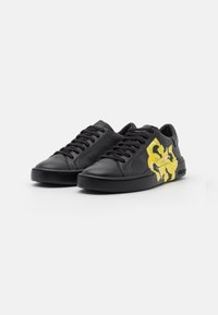 Black low-top sneakers with black laces featuring a yellow abstract design on the side and a thick black sole, angled right on white background.