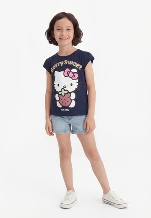 Dziewczynka stoi uśmiechnięta, ubrana w granatowy t-shirt Hello Kitty z napisem „Berry Sweet”, jasne dżinsowe szorty i białe trampki na białym tle.