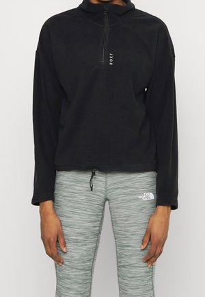 Personne portant un haut polaire Roxy noir avec fermeture éclair sur la moitié et des leggings gris chinés The North Face avec taille à cordon de serrage.