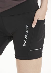 Mustat urheilushortsit, jotka on tehty venyvästä materiaalista. Shortsien vasemmalla puolella on valkoinen "ENDURANCE"-logo ja taskussa heijastinnauha.