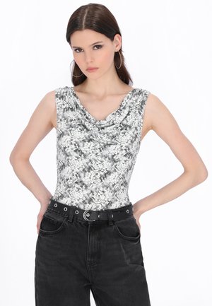 Tânără cu păr drept și castaniu, purtând cercei mari tip cerc, un top cu imprimeu floral alb-negru fără mâneci, blugi negri cu talie înaltă și o curea.