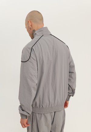 Mann mit Tätowierungen auf Kopf und Hals, der einen lockeren grauen Trainingsanzug mit schwarzer Paspelierung trägt, von hinten vor einem einfarbigen Hintergrund gesehen.