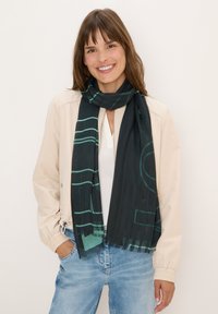 Zwarte sjaal met groene geometrische patronen, lichtgewicht materiaal, franjes aan de randen, gedragen met een beige jasje en een witte top, casual gestyled.