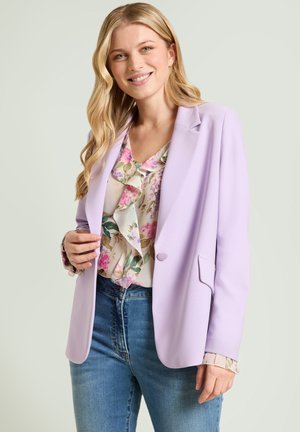 Jonge vrouw die lacht, gekleed in een lichtpaarse blazer over een bloemrijk, gerimpeld blouse en blauwe jeans, staand tegen een effen achtergrond.