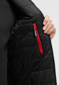 Interno di un giubbotto puffer nero, caratterizzato da una evidente tasca con zip rossa. Il tessuto ha una texture liscia con sezioni trapuntate per l'isolamento.