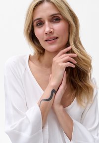 Weißes Seidentop mit langen Ärmeln, getragen von einer Person. Bemerkenswerter schwarzer Schlangentattoo auf dem inneren Unterarm, lässige Pose mit Händen im Gesicht.