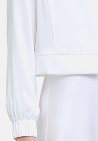 Blouse blanche courte en tissu lisse et léger. Présente des poignets froncés et un design épuré et minimaliste avec un subtil éclat.