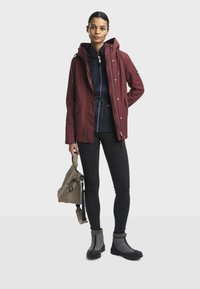 Veste imperméable rouge avec capuche, associée à un pull marine, un legging gris et des bottines noires. Un sac beige est tenu dans une main.