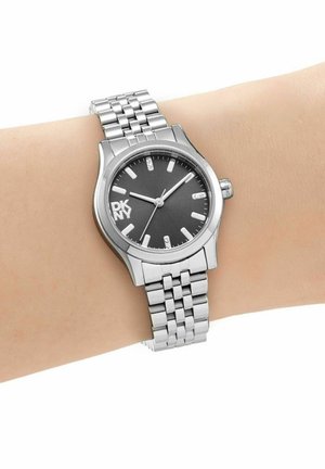 Montre-bracelet en métal argenté avec cadran noir, aiguilles blanches et logo DKNY portée au poignet gauche indiquant l'heure 1:10:35.