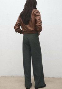 Brun skinn-bomberjacka med ribbade muddar och fåll, kombinerad med vidbyxade mörkgrå byxor. Mjuk textur, minimalistisk design.