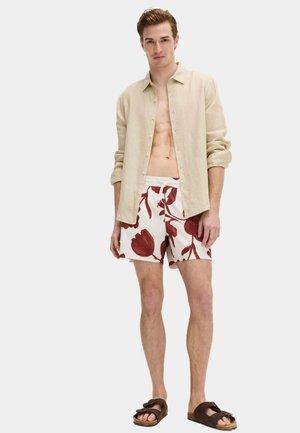 Uomo che indossa una camicia beige a maniche lunghe sbottonata, pantaloncini bianchi con grandi stampe floreali rosse e sandali marrone scuro, in piedi davanti a uno sfondo semplice.