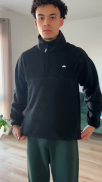 Musta fleece-pulloveri, jossa korkea kaulus, puolipitkä vetoketju edessä ja rintataskussa logo. Siinä on sileä pinta ja rento istuvuus.