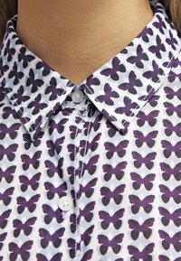 Skjorta med knäppning framtill, i vitt tyg med ett mönster av lila fjärilar som upprepas. Kragen är styv och har en knäppning med en enda knapp.