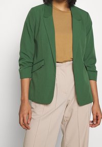 Green blazer with a tailored fit, featuring a single-button closure, shoulder pads, and two side pockets, paired with beige trousers.