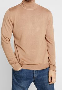 Beige rullekrave trøje lavet af et blødt, let materiale. Har lange ærmer og en ribbet nederste kant, bæres med blå jeans.