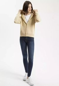 Beżowa bluza z zamkiem i regulowanym kapturem, z kieszeniami z przodu i miękką fakturą, zestawiona z ciemnoniebieskimi skinny jeansami i białymi sneakersami.