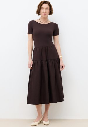 Kobieta stoi, ubrana w krótką sukienkę midi z ciemnobrązowego materiału z falbanami, beżowe niskie obcasy, złoty naszyjnik łańcuszkowy i złoty zegarek na nadgarstku.