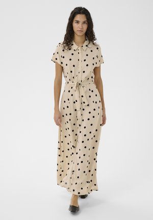 Kobieta w beżowej sukience maxi w czarne kropki, z przodem na guziki, przewiązana w talii, w czarnych płaskich butach, stojąca na białym tle.