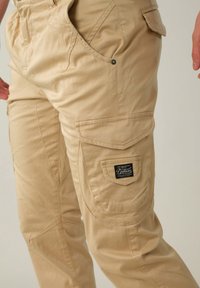 Beige cargobroek gemaakt van duurzaam materiaal, met meerdere zakken, een label met "Deeluxe" en een ontspannen pasvorm.