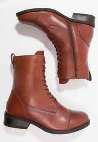 Botas de tobillo de cuero marrón con punta redondeada, cremallera lateral, detalle de cordones y un tacón bajo y rectangular. Textura suave con diseño minimalista.
