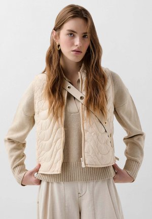 Beige gewatteerd vest met een doorstikte textuur, voorzien van een zijrits, drukknoopsluiting en een hoge kraag. Onder het vest wordt een gebreide beige trui gedragen.