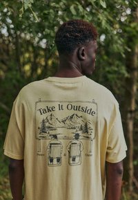 T-shirt beige clair oversize avec un motif graphique représentant des montagnes, des arbres et deux personnages assis, accompagné du texte « Prenez l'air ». 