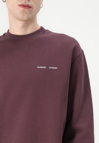 Felpa bordeaux con scollatura a costine, caratterizzata da testo bianco "SAMØSÆ SAMSØE" sul petto. Tessuto morbido, vestibilità rilassata e maniche lunghe.