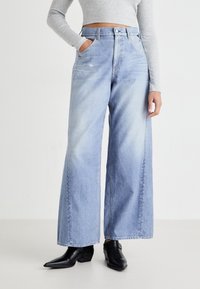 Svetlo modri široki jeans iz denima z visokim pasom, ki imajo dve sprednji žepni in subtilne podrobnosti zbledelosti. V kombinaciji s črnimi gležnjački.